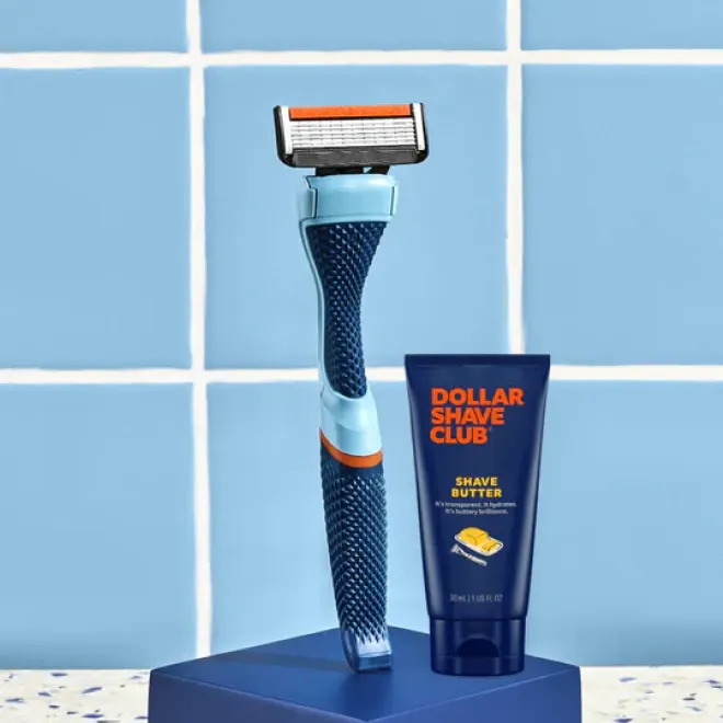 Blue and orange razor with Dollar Shave Club shave butter cream on a blue stand against blue tile.